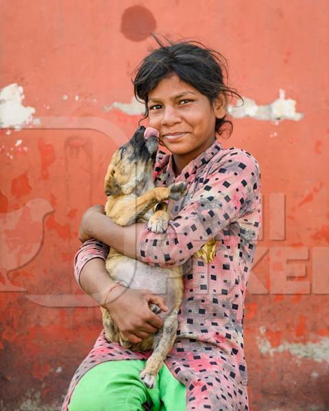 Portrait of girl holding Indian street or stray puppy dog with orange wall background, Jaipur, India, 2022